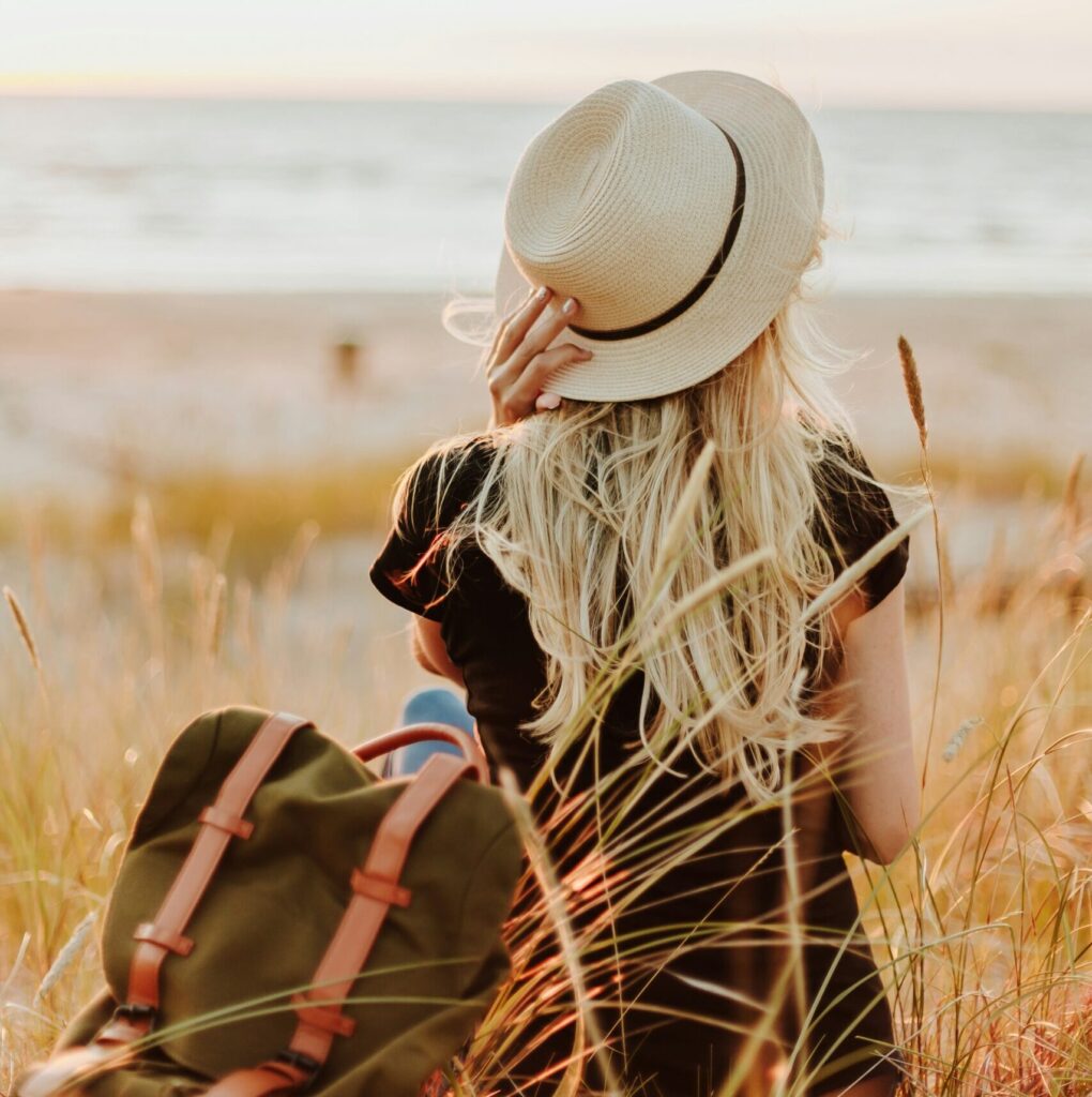 Frau mit Sonnenhut und Rucksack sitzt entspannt in der Natur – bewusster Urlaub und langsameres Reisen